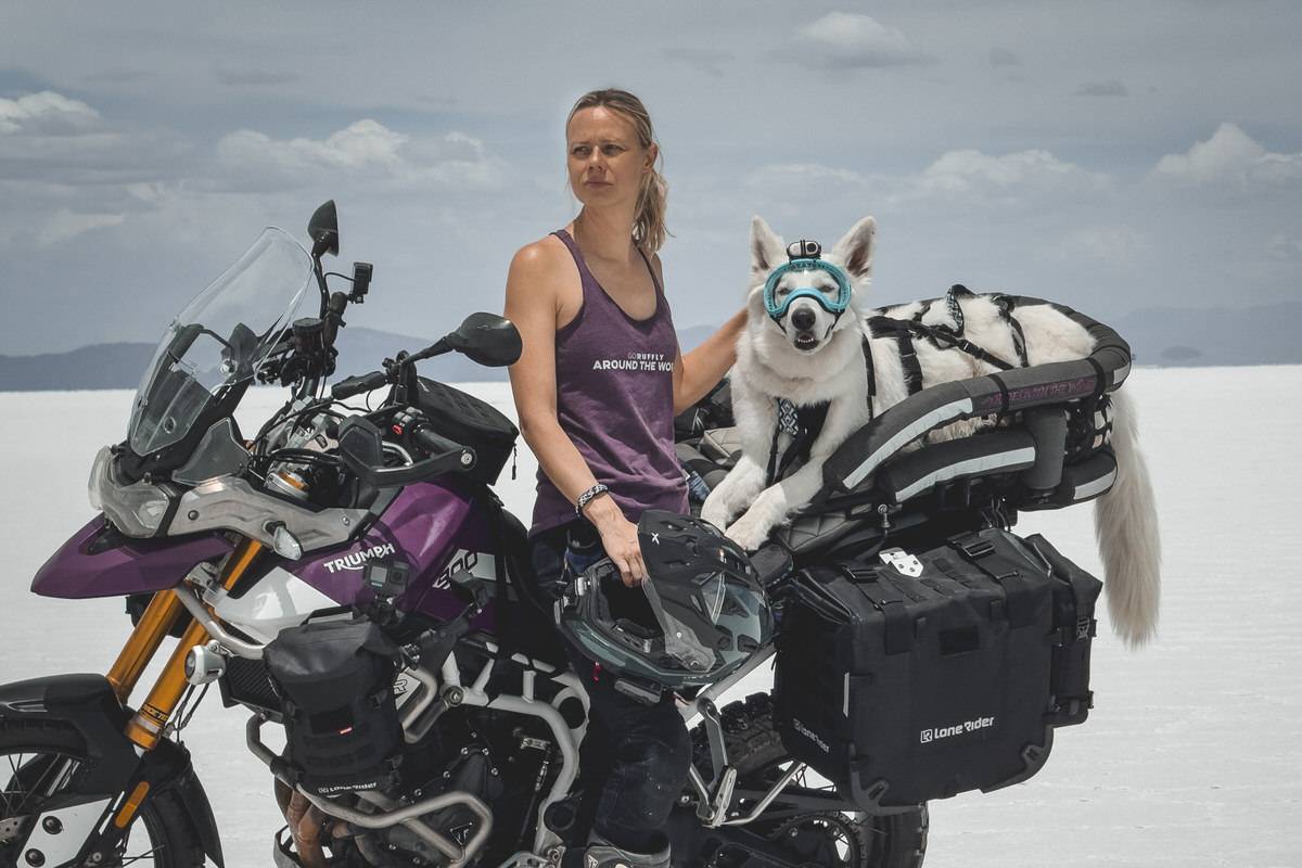 Blonde woman leans on motorcycle with white dog in carrier on salt flats