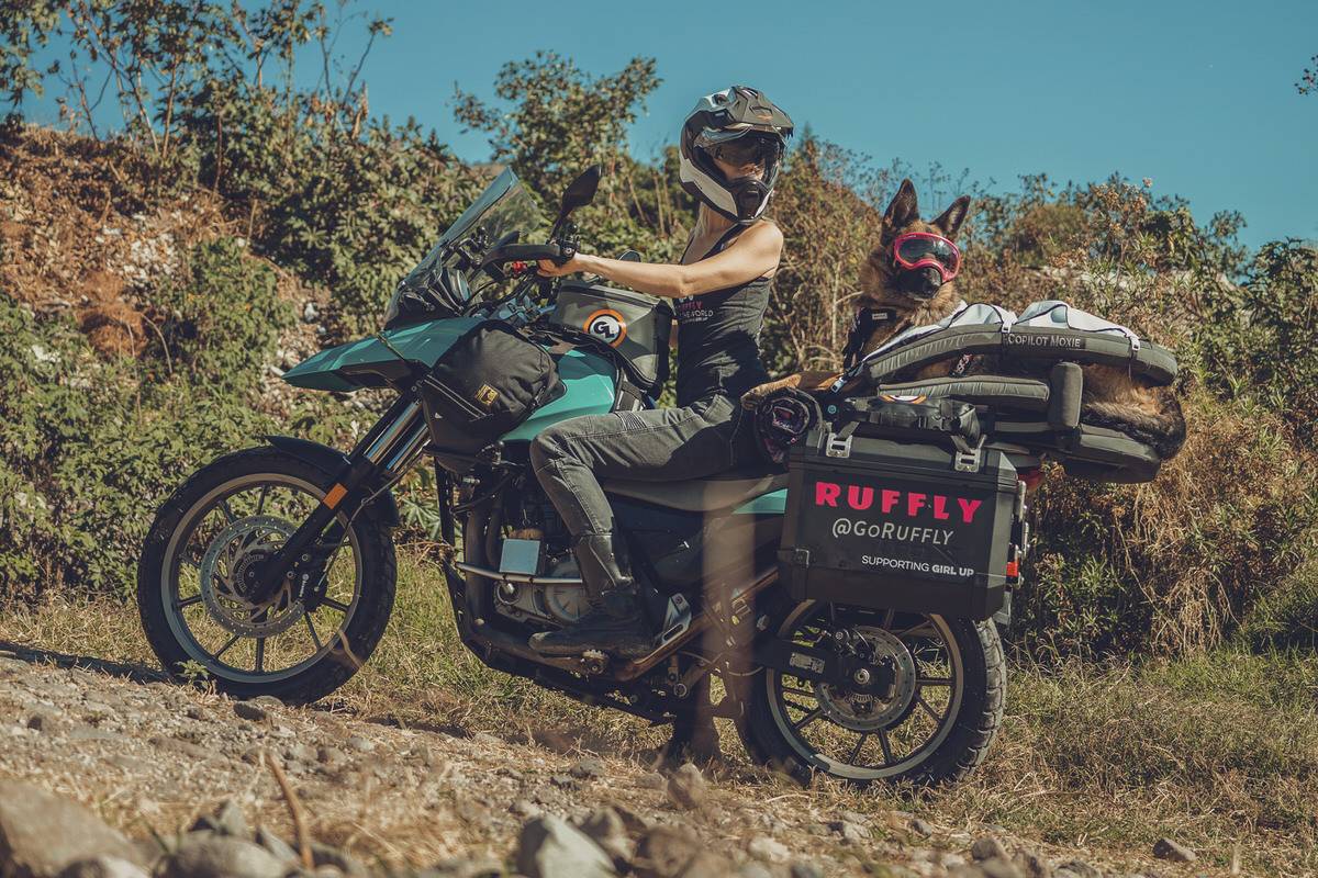 Woman wearing riding helmet with sunshade beside a German Shepherd wearing goggles and a sunshield in a motorcycle dog carrier