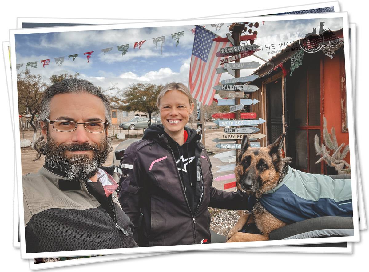 Man, woman, and German Shepherd dog stand beside American flag and sign post