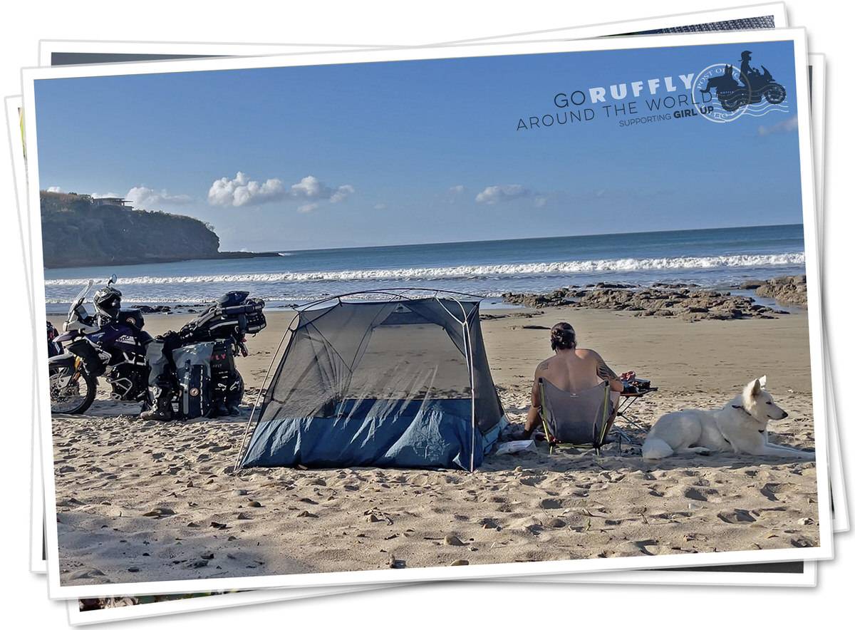 Man sits on the beach beside a white dog, a tent, and a motorcycle