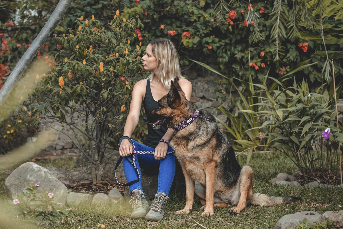Everyday But Never Ordinary Blonde woman holds craftsman leash and sits in a garden beside a german shepherd wear wearing a matching collar