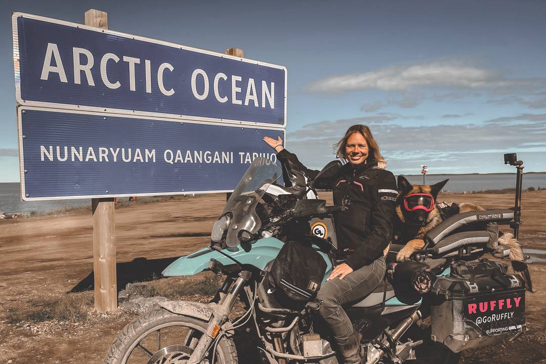 Woman points to Arctic Ocean sign with German shepherd on her motorcycle
