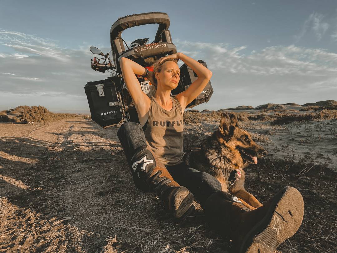 Woman sits against motorcycle on sand road beside German shepherd dog