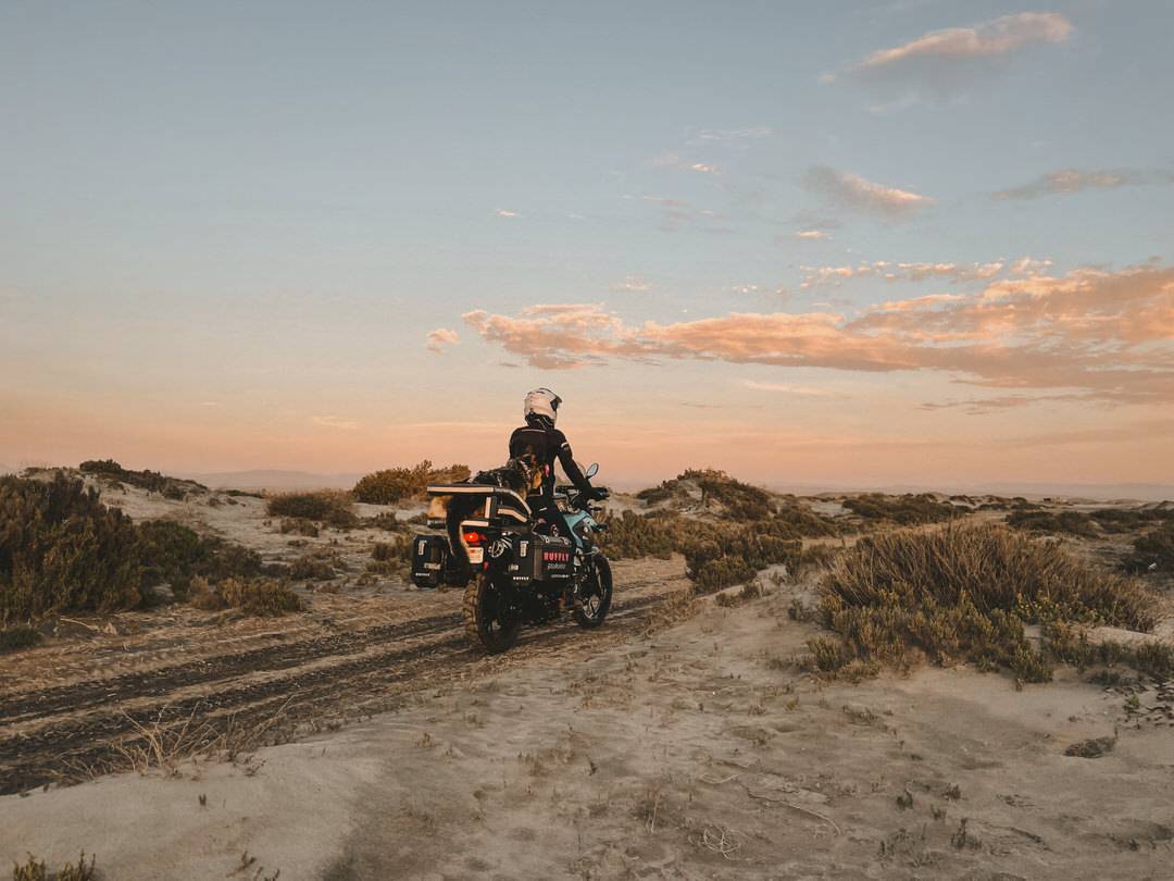 Woman rides motorcycle through sand with German shepherd dog on the back