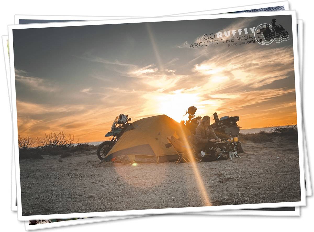 Postcard of a woman camping beside two motorcycles by the sea at sunset