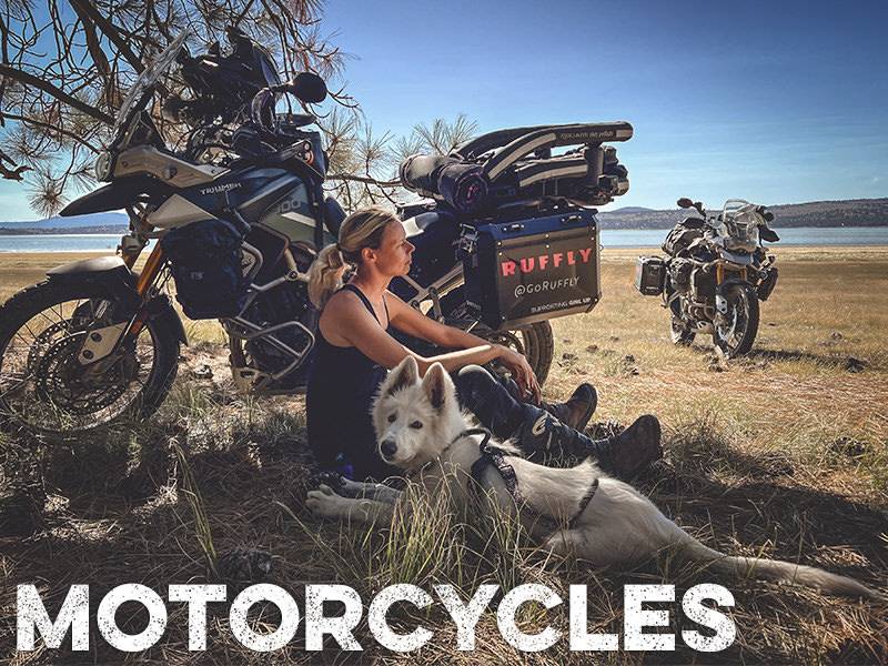 Woman reclines against adventure motorcycle with a white dog in the shade with overlaid text: "Motorcycles"