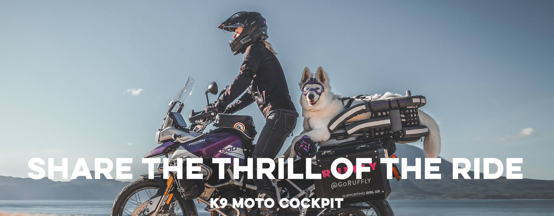 HEADER PHONE 2·56x1 20210607 Woman stands while riding an adventure motorcycle off-road with a white shepherd dog on the back