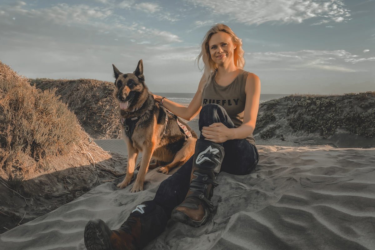 The Bond Blonde woman sits on sand dune next to German Shepherd