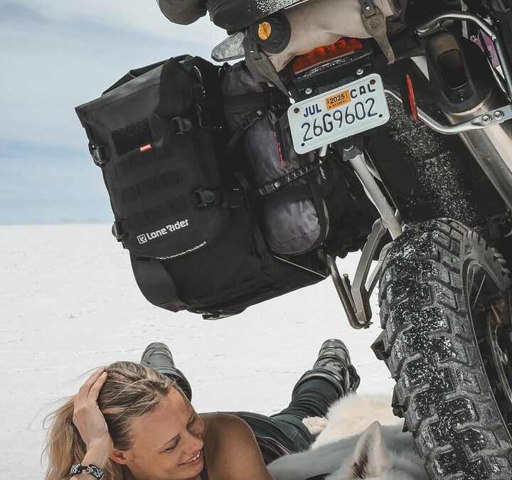 Woman and white dog lay on dog travel bed on white ground under the shade of a motorcycle